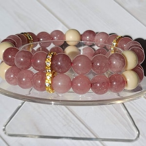 Könnte beinhalten: Zwei übereinanderliegende Armbänder aus rosa und weißen Perlen mit einem goldenen Band aus Strasssteinen. Die Armbänder befinden sich auf einem durchsichtigen Ständer.