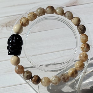 May include: A brown and white beaded bracelet with a black charm. The bracelet is on a clear display stand.