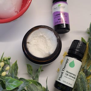 May include: A red bowl filled with white scrub, a jar of white cream, and two small essential oil bottles. One bottle is labelled "Lavender" and the other "Rosemary 1,8-Cineole". Green leafy sprigs and small yellow flowers surround the products.