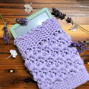 Puede incluir: Una funda de ganchillo de color morado claro con un patrón de puntos de estrella texturizados, que contiene parcialmente un lector electrónico de color verde claro. La funda está rodeada de lavanda seca y flores blancas.