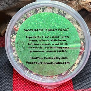May include: A round, clear plastic container filled with a dried food mix labeled Sasquatch Turkey Feast. The green glittery label lists ingredients including turkey, collards, beans, squash, cranberries, strawberries, and coconut.