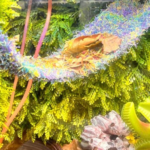 May include: A terrarium scene featuring a colorful hammock made of blue, purple, and yellow fibers, with a brown leaf inside. The scene is surrounded by vibrant green artificial plants and pink support structures.