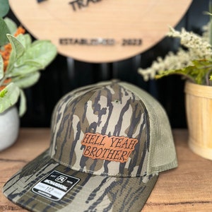 May include: A camouflage trucker hat with a brown leather patch that says "Hell Yeah Brother!" The hat is made by Richardson and has a size 112 tag.