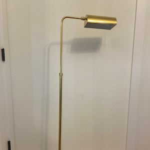 May include: A gold-toned floor lamp with a rectangular shade. The lamp has a slender, adjustable pole and a rounded base. The shade is black on the bottom. The lamp is on a wooden floor.