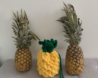May include: A yellow and green crocheted pineapple-shaped bag with a green strap. Two real pineapples are on either side of the bag.