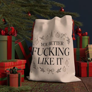 May include: A cream-colored fabric drawstring gift bag featuring the black text "YOU BETTER FUCKING LIKE IT" centered on the front, surrounded by a decorative circular pattern of line-art Christmas ornaments, bows, and pine branches.