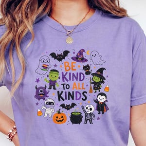 May include: A light purple short-sleeve t-shirt featuring a graphic print of various Halloween characters including a ghost, mummy, vampire, and witch. The text "BE KIND TO ALL KINDS" is printed in the center in purple and orange lettering.