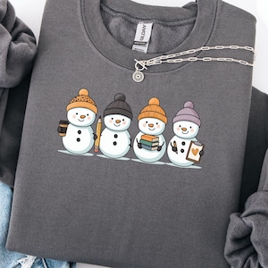 May include: Dark grey sweatshirt with a graphic of four snowmen wearing hats and holding a pencil, books, and a notepad. The snowmen are on a white line. A silver necklace and a pair of blue jeans are also in the image.