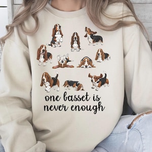 May include: A cream-coloured sweatshirt displaying cartoon basset hounds in different poses. The text "one basset is never enough" is printed in black script. The sweatshirt is a casual, comfortable style, suitable for everyday wear.