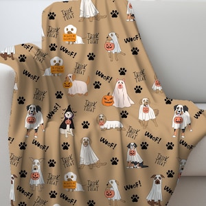 May include: A tan throw blanket with a repeating pattern of dogs dressed in Halloween costumes. The dogs are wearing ghost costumes, pumpkin costumes, and trick or treat signs. The blanket has black paw prints and the words "Woof!" and "Trick or Treat" repeated throughout.