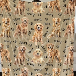 May include: A tan fleece blanket with a repeating pattern of golden retriever dogs and the name "Abbey" printed on it.