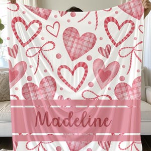 Peut inclure: Une couverture blanche avec un motif de cœurs roses, de nœuds et de pois. La couverture comporte une bande rose avec le nom "Madeline" imprimé dessus. Les cœurs ont des motifs unis, rayés et à carreaux.
