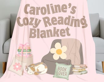 Peut inclure: Une couverture rose clair avec le texte "Caroline's Cozy Reading Blanket" en marron. La couverture présente des illustrations d'un coin lecture avec un chat, des livres et une fleur. La couverture est tenue par une personne.