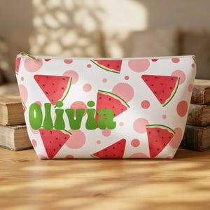 Può includere: Un beauty case bianco con chiusura a zip, con un motivo a fette di anguria e pois rosa. Il nome "Olivia" è stampato in verde, aggiungendo un tocco personalizzato a questo accessorio a tema estivo.