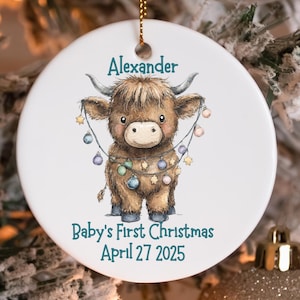 May include: A round white Christmas ornament with a Highland cow design. The cow is depicted in brown with a light brown face, adorned with a string of colorful lights. The ornament includes the name "Alexander" and the text "Baby's First Christmas April 27 2025".