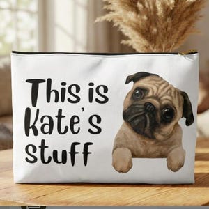 May include: White zippered pouch with the text "This is Kate's stuff" in black. A watercolor illustration of a pug dog is on the right side. The pouch is on a wooden surface.