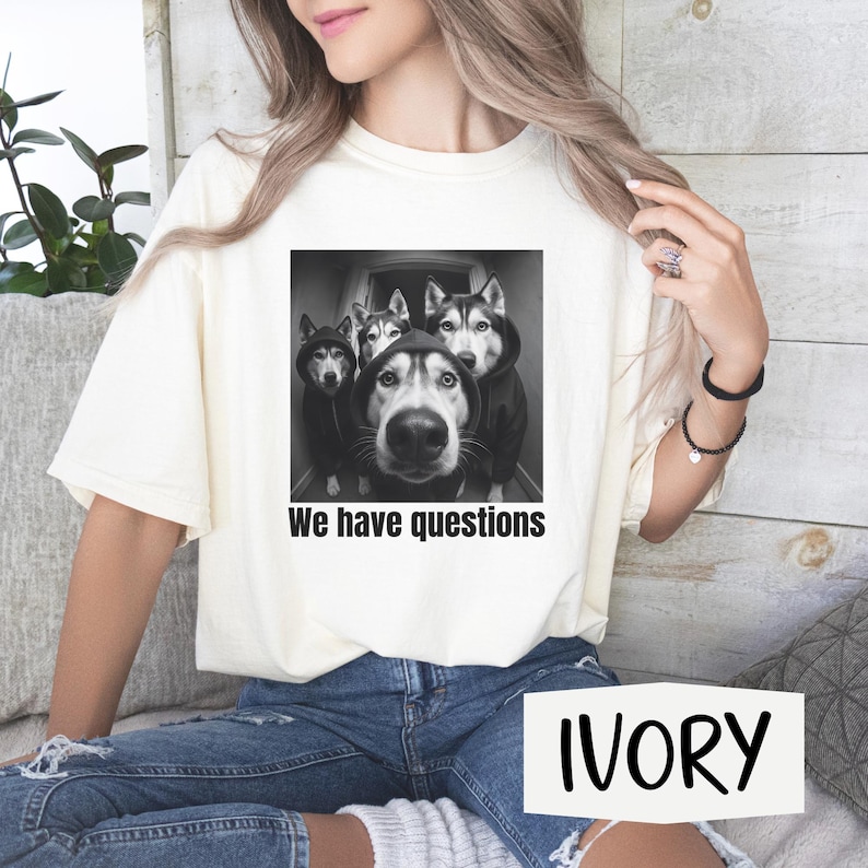 May include: Ivory t-shirt with a black and white graphic of four Huskies in hoodies, and the text "We have questions". The shirt is a relaxed fit. The word "IVORY" is printed on the bottom right.