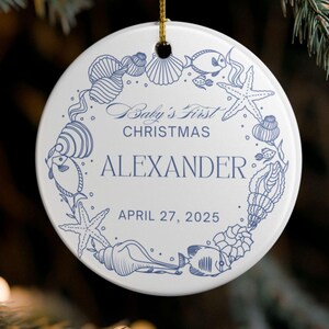 Może przedstawiać: Biała ceramiczna ozdoba świąteczna z niebieskim wzorem muszli i życia wodnego. Tekst brzmi "A Baby's First Christmas" i "Alexander, April 27, 2025". Złoty sznurek jest przymocowany do zawieszenia.