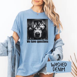 May include: Pale blue t-shirt with a black and white graphic of four huskies in hoodies, with the text "We have questions". The shirt is made of a soft material and is a washed denim colour.