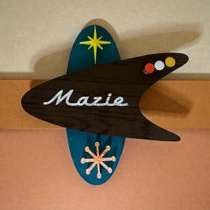 May include: A retro-style wooden sign with the name "Mazie" in white script. The sign features a teal oval shape with a yellow star and an orange starburst. A dark brown boomerang shape has the name "Mazie" and three colored dots.