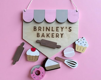 May include: Wooden sign with the text "BRINLEY'S BAKERY" and a pink and gray awning design. Includes wooden cutouts of a rolling pin, cupcake, donut, macaron, cake slice, and whisk. The sign hangs from a white string.