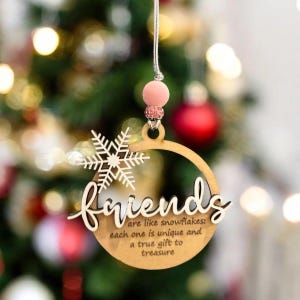 May include: A wooden Christmas ornament with the word "friends" and a snowflake design. The ornament has a light pink bead and silver string for hanging. Text on the ornament reads "are like snowflakes: each one is unique and a true gift to treasure."