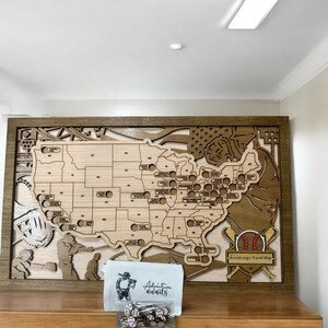 May include: A wooden baseball travel map of the United States, with a dark brown frame. The map features cutouts for baseball stadiums and includes a small bag with baseball-themed pins. The text "Adventure Awaits" is visible.