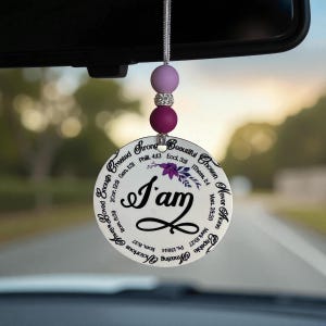 May include: A round car charm with the words "I am" in a cursive font, surrounded by inspirational words and Bible verses. The charm is white with purple floral accents and is suspended by a silver cord and purple beads.
