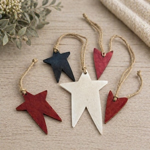 May include: A collection of rustic wooden star and heart ornaments in red, white, and navy blue. Each ornament is strung with natural twine. The ornaments are a patriotic theme and are suitable for holiday or everyday decor.