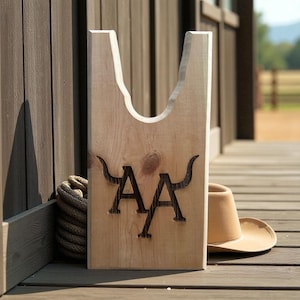 May include: A light-colored wooden boot jack with a cutout design and the letters "AA" in a dark color. A tan cowboy hat and a coiled rope are next to the boot jack. The boot jack is on a wooden deck.