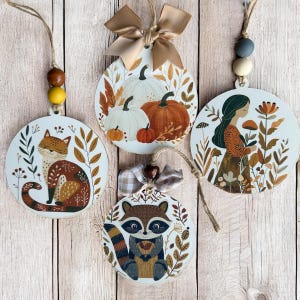 May include: Four round, wooden ornaments with fall-themed designs. One features a fox, another pumpkins, a third a woman in a field, and the last a raccoon. Each ornament has a jute string and decorative beads or a bow for hanging.