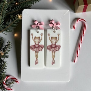 May include: A pair of white rectangular earrings featuring a ballerina frog design. Each earring has a pink bow at the top and a frog in a pink tutu. A candy cane and a gift box are in the background.