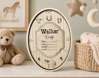 May include: Oval wooden baby announcement sign with a Western theme. The sign features the name "Walker Cody" and spaces for date, time, weight, and length. Decorative elements include a horseshoe, cowboy hat, and a horse.