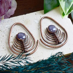Puede incluir: Dos pendientes en forma de lágrima de color cobre con una piedra púrpura central y un diseño detallado en forma de costillas. Los pendientes se exhiben sobre una superficie clara, con flores y vegetación de fondo.