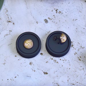 May include: Two dark gray stud earrings with a circular design. Each earring features a light pink center with gold leaf accents. One earring is displayed on its back, the other is upright.