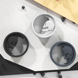 Può includere: Tre elementi decorativi circolari con un teschio all'interno di una luna crescente. Uno è nero, uno bianco e l'altro grigio. Sono disposti su una superficie bianca e grigia con forme geometriche nere e gialle.