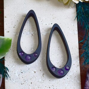 Può includere: Un paio di orecchini a goccia grigio scuro. Ogni orecchino presenta tre pietre preziose rotonde viola nella parte inferiore. Sono esposti su una superficie strutturata bianco sporco, con accenni di verde e fiori sullo sfondo.