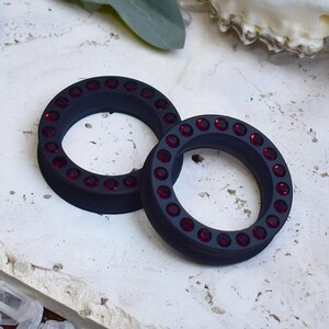 Può includere: Due dilatatori per orecchie neri, circolari, con un anello di piccole gemme rosse. I dilatatori sono in materiale opaco e sono esposti su una superficie chiara. Gioielli per piercing all'orecchio allungati.