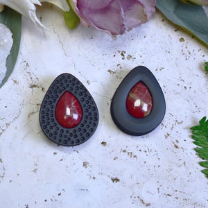 May include: Two teardrop-shaped earrings. One earring has a black border with small circular details around a red stone. The other earring has a smooth black border around a red stone. The earrings are displayed on a white surface with floral accents.