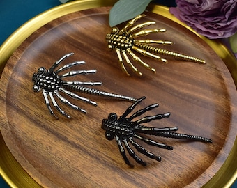 May include: Three metal alien-like creature brooches in gold, silver, and black. The brooches are shaped like a spider with long legs and a segmented body.