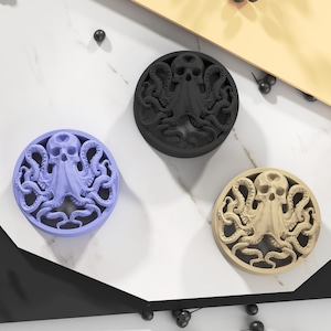 Può includere: Tre oggetti decorativi rotondi con un design a teschio e polpo. Uno è lavanda, uno è nero e l'altro è beige. Gli oggetti sono su una superficie bianca e nera con un accento giallo.
