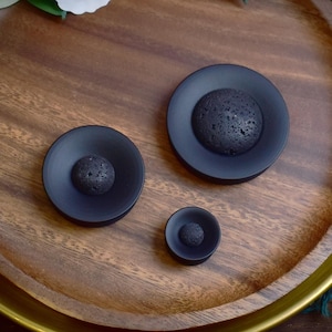 Può includere: Tre tappi neri circolari con un centro testurizzato grigio scuro, esposti su un vassoio di legno. I tappi variano di dimensioni, il più grande misura circa 2,5 cm di diametro. I tappi hanno un design minimalista.