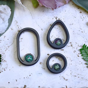 May include: Three dark gray metal pendants in different shapes: an oval, a teardrop, and a circle. Each pendant features a small, round, green and purple stone at the center. The pendants are displayed on a textured, off-white surface.