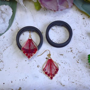 May include: Gold-toned, teardrop-shaped earrings with red thread and red gemstone accents. The earrings are displayed with two black circular objects, possibly earring holders, against a textured, off-white surface. The earrings have a delicate, handcrafted appearance.