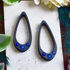 Può includere: Due orecchini a goccia grigio scuro con tre accenti sferici blu. Gli orecchini sono esposti su una superficie strutturata bianco sporco. Design moderno e minimalista.