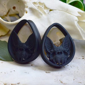 Può includere: Due dilatatori per orecchie neri a forma di goccia con un design a pipistrello. Il pipistrello è intagliato in dettaglio, con ali e lineamenti del viso visibili. I dilatatori sono posti su uno sfondo sfocato.