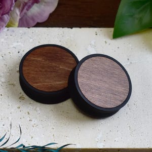 May include: Two round wooden plugs with a dark brown wood grain design and black rims. The plugs are displayed on a light-colored, textured surface. The plugs are likely for ear piercings.