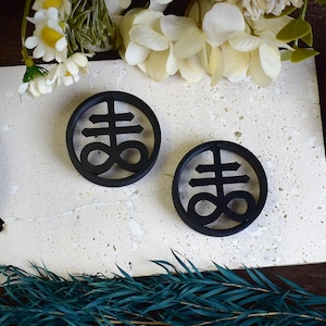 May include: A pair of black circular earrings featuring a raised, stylised symbol of a cross with an infinity symbol at the base.