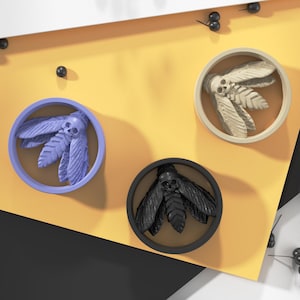 Può includere: Tre sottobicchieri rotondi e decorativi con un design a forma di teschio e ali. I sottobicchieri sono di colore nero, bianco e viola.