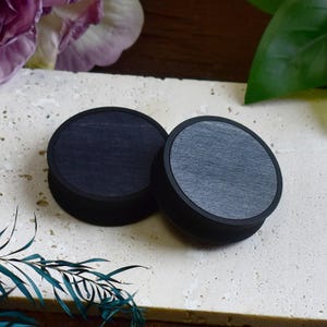 Può includere: Due tappi in legno neri e grigi. I tappi hanno un design circolare con un centro a venature di legno grigio scuro e un bordo nero. I tappi sono esposti su una superficie di colore chiaro.